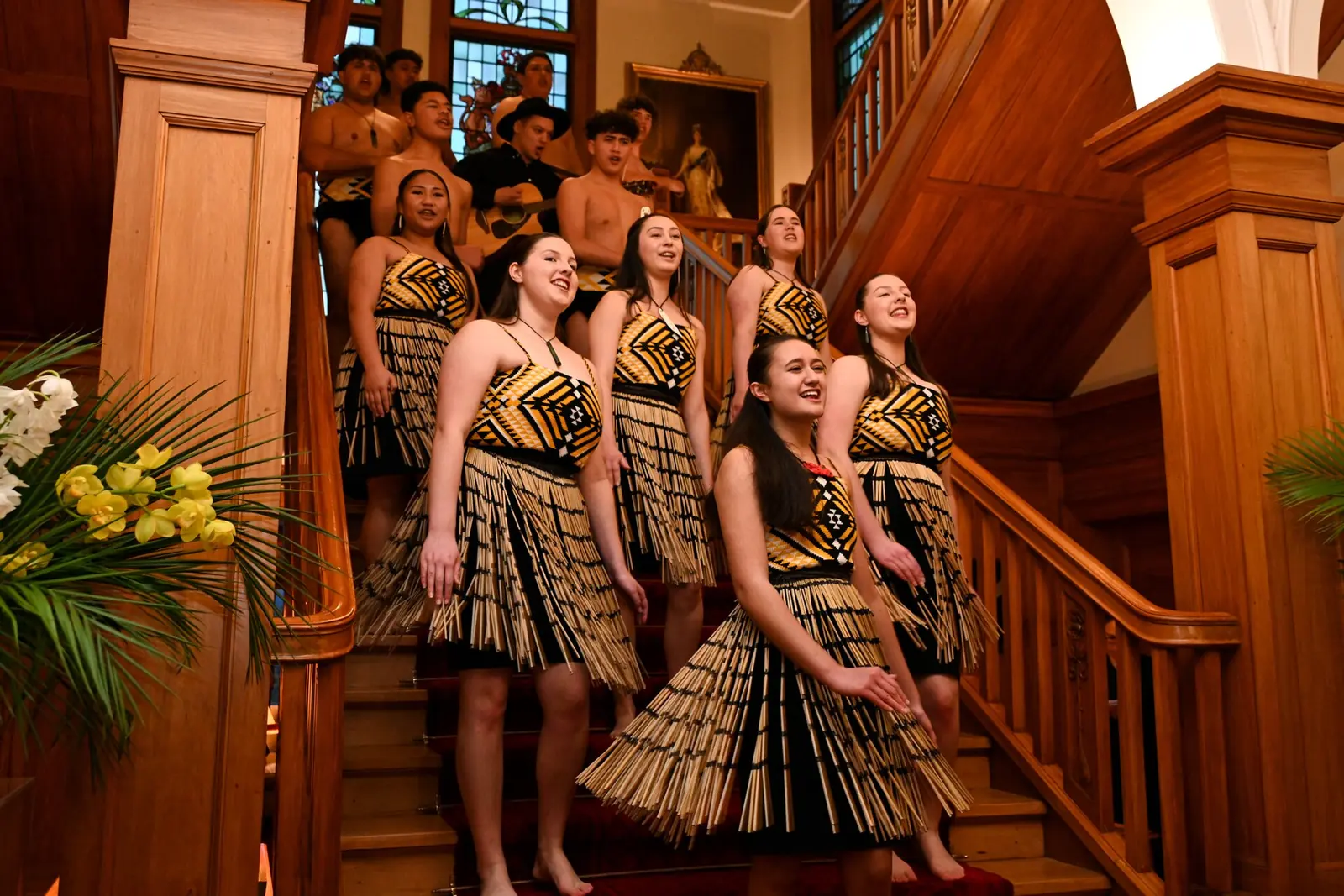 Eine Kapa-Haka-Gruppe in gewebten Flachsröcken, die auf einer Holztreppe singt, mit weiteren Darstellern dahinter (nicht im Auckland Museum)