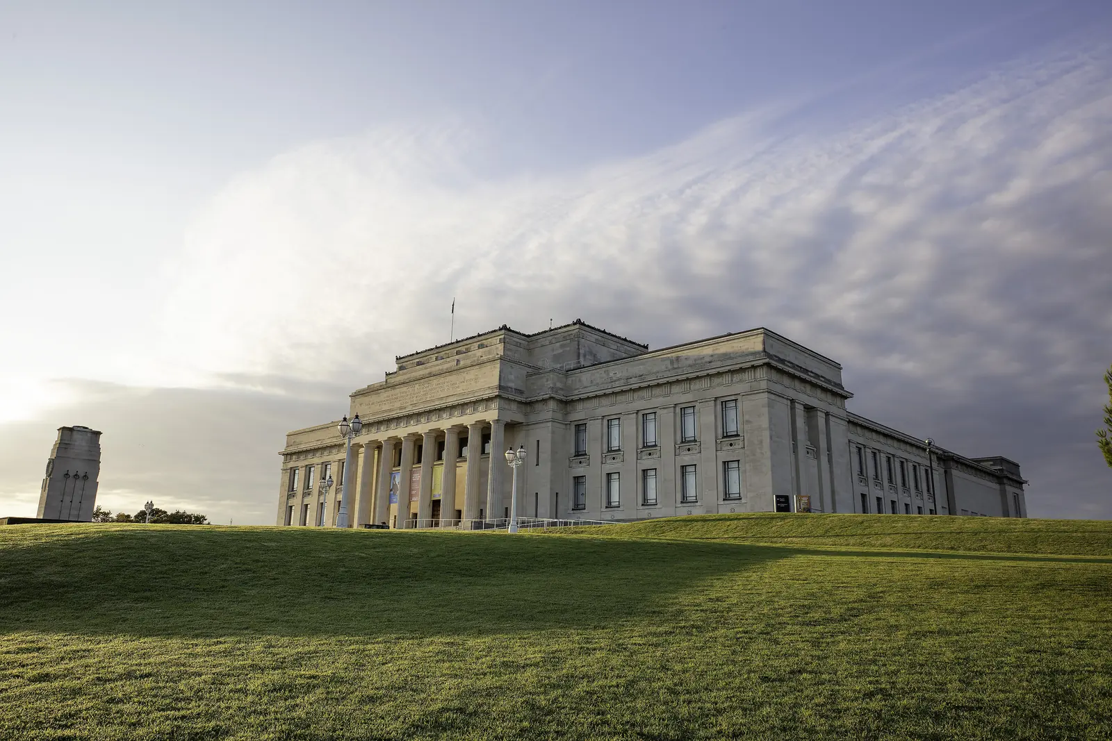 Das Auckland War Memorial Museum auf seinem grasbewachsenen Hügel in der Abenddämmerung, mit dem von der tief stehenden Sonne beleuchteten Säulenportikus und dem Kenotaph-Turm links