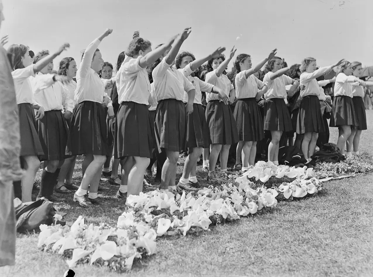 Eine Gruppe von Schulmädchen in weißen Blusen und dunklen Röcken, die in Reihen auf dem Gras stehen und einen Arm erhoben haben, mit Liliensträußen vor sich auf dem Boden