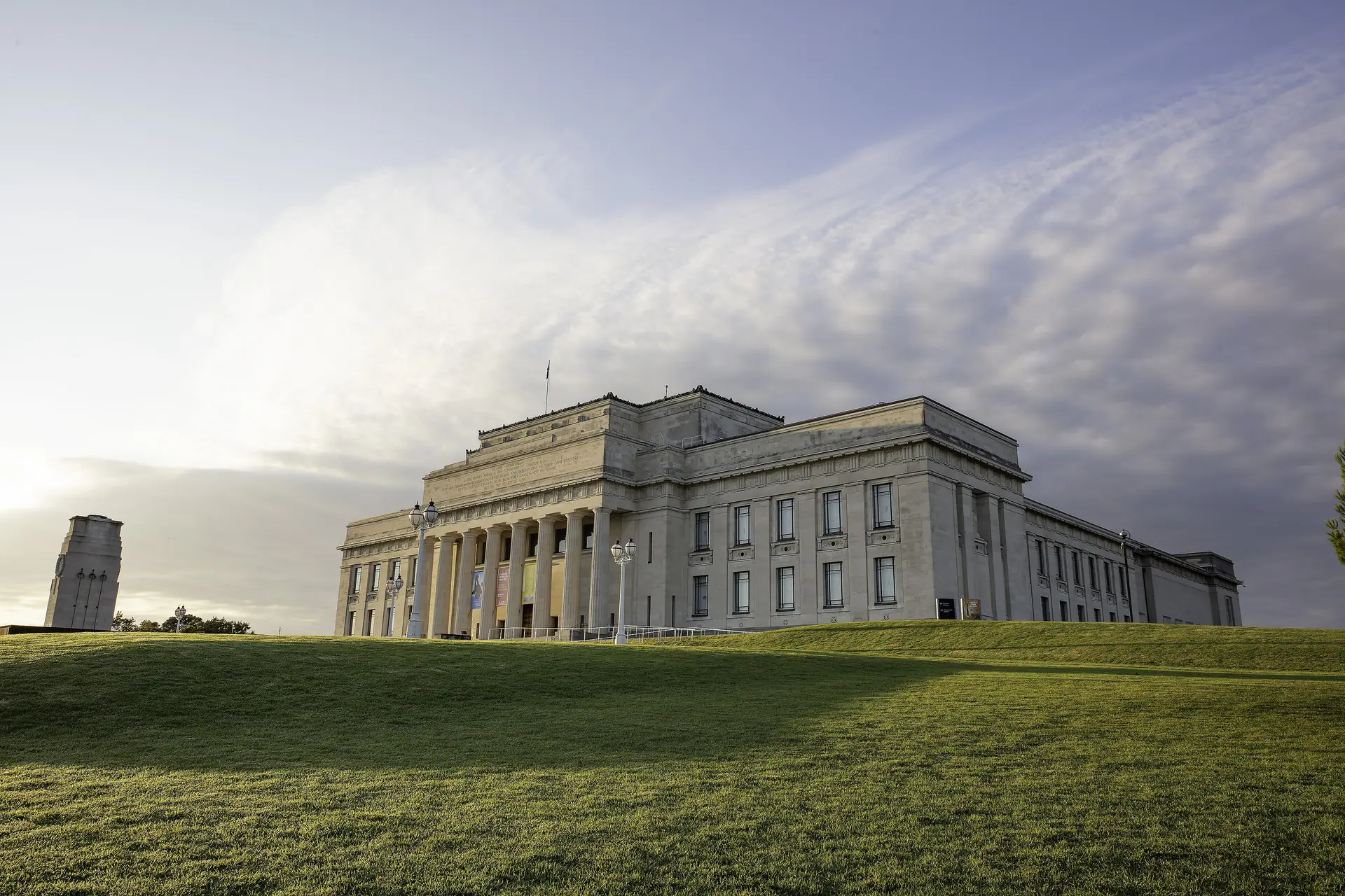 Das Auckland War Memorial Museum in der Abenddämmerung, seine neoklassizistische Säulenfront von tiefer Sonne beschienen, über einem grasbewachsenen Hang im Auckland Domain